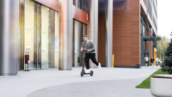 Attractive young man riding contemporary kick scooter at cityscape background.