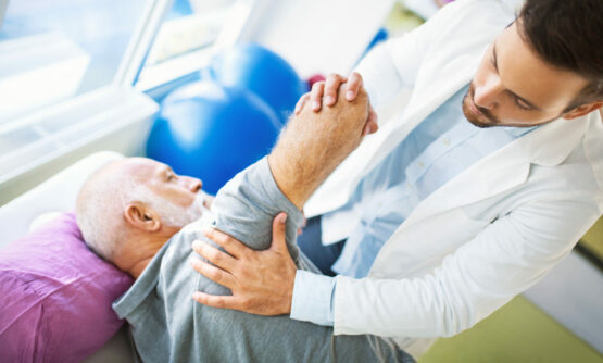 Closeup top view of an early 60's senior gentleman having some shoulder pain. He's at doctor's office having medical examination by a male doctor. The doctor is touching the sensitive area and trying to determine the cause of pain.