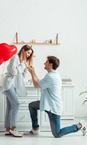 happy man standing on knee while doing marriage proposal to excited woman at home